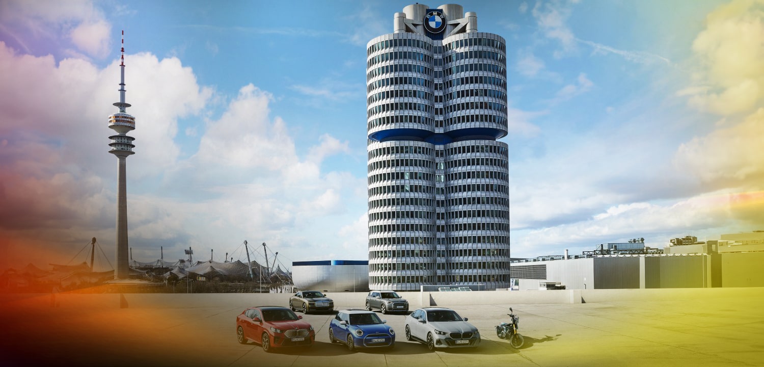 Vor einem großen Turm mit BMW-Logo stehen nebeneinander aufgereiht ein BMW, ein MINI, ein weiterer BMW und ein BMW Motorrad. Dahinter stehen ein weiterer MINI und ein Rolls-Royce.